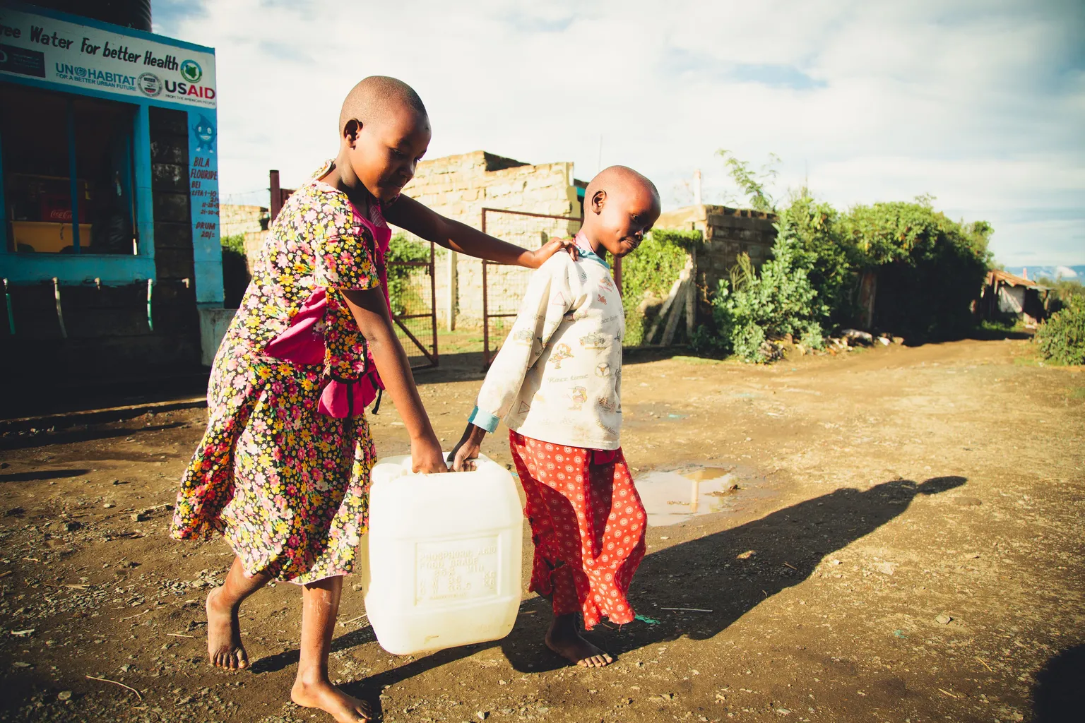 Two children walk on a dirt road, sharing the task of carrying a large water container under a bright sky, with buildings and greenery in the background.