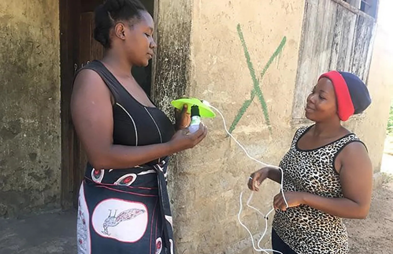 Two women stand outside a building, using a portable solar-powered device. One holds the device while the other holds connected wires.
